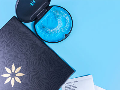 The image shows a blue toothbrush with a clear case containing cleaning crystals placed on top of a dark-colored book or box with a floral pattern, next to an open blue package that appears to be a dental product.