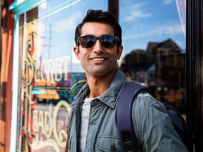 A smiling man wearing sunglasses and a backpack stands confidently outside a storefront with a neon sign.